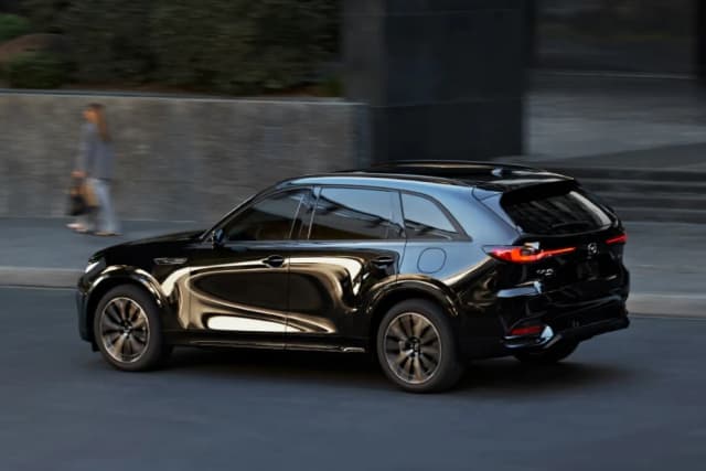 Mazda CX-70 in black driving past a building, rear three-quarter view