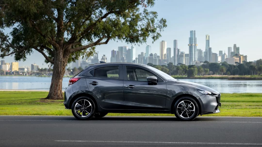 Mazda2 hatch in grey parked by a lake with a city skyline behind, side view