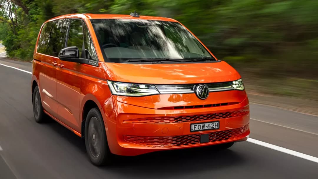 Orange and white Volkswagen Multivan driving on a forest road, front three-quarter view
