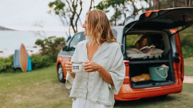 Woman with a coffee beside an orange Volkswagen Multivan with the tailgate open at the beach