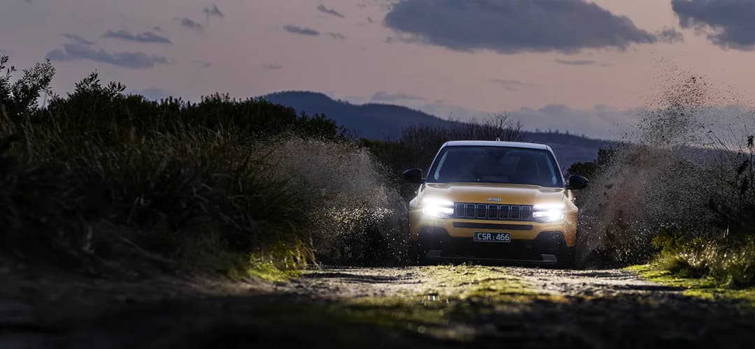 Jeep Avenger in yellow charging along a dirt track at dusk with headlights on